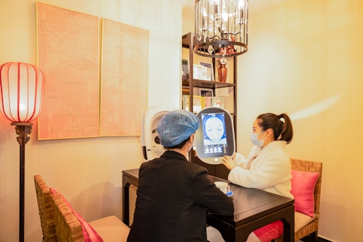 Two people are seated at a small table in a well-lit room, engaging with a digital screen that displays facial imagery. One person, wearing a mask and a white coat, admires the screen while the other, in darker attire with a blue hair cover, observes. The surroundings feature warm lighting, a decorative lamp, artworks on the wall, and bookshelves filled with books or decorations.