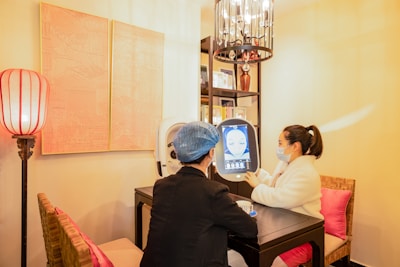 Two people are seated at a small table in a well-lit room, engaging with a digital screen that displays facial imagery. One person, wearing a mask and a white coat, admires the screen while the other, in darker attire with a blue hair cover, observes. The surroundings feature warm lighting, a decorative lamp, artworks on the wall, and bookshelves filled with books or decorations.