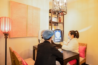 Two people are seated at a small table in a well-lit room, engaging with a digital screen that displays facial imagery. One person, wearing a mask and a white coat, admires the screen while the other, in darker attire with a blue hair cover, observes. The surroundings feature warm lighting, a decorative lamp, artworks on the wall, and bookshelves filled with books or decorations.