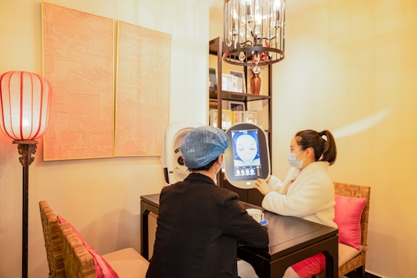 Two people are seated at a small table in a well-lit room, engaging with a digital screen that displays facial imagery. One person, wearing a mask and a white coat, admires the screen while the other, in darker attire with a blue hair cover, observes. The surroundings feature warm lighting, a decorative lamp, artworks on the wall, and bookshelves filled with books or decorations.