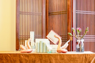 A neatly arranged tabletop display featuring cylindrical cosmetic bottles, two artistic sculptures resembling swans, and a glass vase with tulips. The setup is placed against a traditional wooden screen with vertical slats.