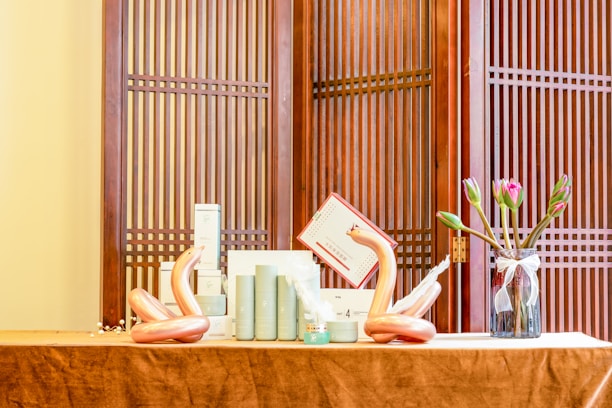 A neatly arranged tabletop display featuring cylindrical cosmetic bottles, two artistic sculptures resembling swans, and a glass vase with tulips. The setup is placed against a traditional wooden screen with vertical slats.