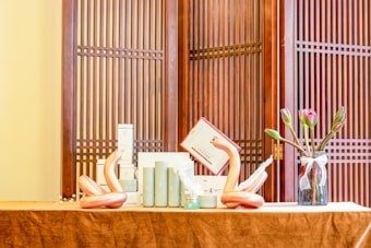 A neatly arranged tabletop display featuring cylindrical cosmetic bottles, two artistic sculptures resembling swans, and a glass vase with tulips. The setup is placed against a traditional wooden screen with vertical slats.