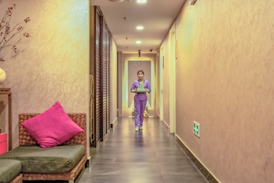 A hallway with soft lighting features a person walking towards the camera, holding a tray. The walls are a warm beige color, and there is a green cushioned bench with a bright pink pillow on the left. Overhead lights illuminate the corridor, and there are doors along the right side.