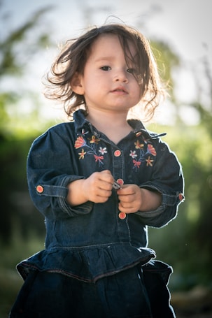 A young child with wavy hair wears a dark blue denim dress with colorful floral embroidery. The child holds an object in their hands and stands outdoors with soft natural lighting. The background appears to be a blurred green landscape, likely indicating a garden or park setting.