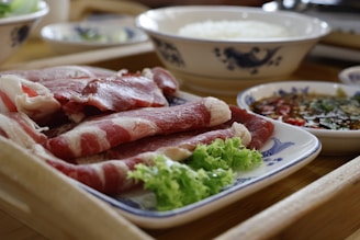 A plate of raw, marbled beef slices is attractively arranged, accompanied by fresh green lettuce. Nearby, a small dish holds a flavorful dipping sauce garnished with herbs and chili pieces, and a bowl of white rice sits in the background.