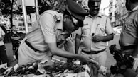 A group of police officers in uniform engage with a child across a table laden with roses. One officer leans forward, smiling and shaking hands with the child, while another holds a rose. The scene takes place on a bustling city street with various people and buildings in the background.