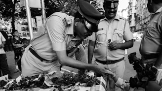 A group of police officers in uniform engage with a child across a table laden with roses. One officer leans forward, smiling and shaking hands with the child, while another holds a rose. The scene takes place on a bustling city street with various people and buildings in the background.
