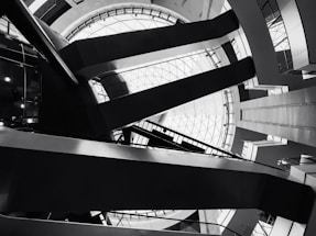 Architectural interior featuring geometric shapes and sharp lines. The design includes a large circular skylight at the top, surrounded by intersecting beams and various levels of walkways. The image is in black and white, emphasizing the contrast and structure.