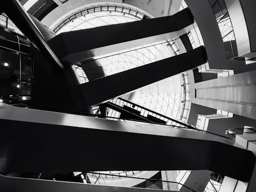 Architectural interior featuring geometric shapes and sharp lines. The design includes a large circular skylight at the top, surrounded by intersecting beams and various levels of walkways. The image is in black and white, emphasizing the contrast and structure.