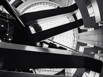 Architectural interior featuring geometric shapes and sharp lines. The design includes a large circular skylight at the top, surrounded by intersecting beams and various levels of walkways. The image is in black and white, emphasizing the contrast and structure.