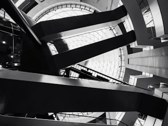 Architectural interior featuring geometric shapes and sharp lines. The design includes a large circular skylight at the top, surrounded by intersecting beams and various levels of walkways. The image is in black and white, emphasizing the contrast and structure.