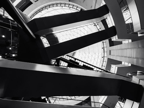 Architectural interior featuring geometric shapes and sharp lines. The design includes a large circular skylight at the top, surrounded by intersecting beams and various levels of walkways. The image is in black and white, emphasizing the contrast and structure.