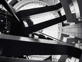 Architectural interior featuring geometric shapes and sharp lines. The design includes a large circular skylight at the top, surrounded by intersecting beams and various levels of walkways. The image is in black and white, emphasizing the contrast and structure.