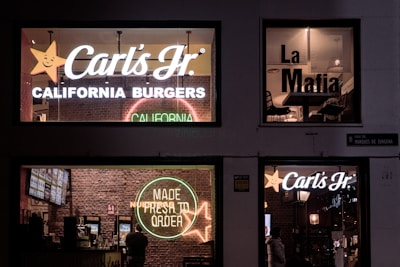 A nighttime scene of a building with illuminated signs for two establishments. The left side features Carl's Jr. with bright, white, and neon signage against a brick wall, displaying 'California Burgers' and 'Made Fresh to Order.' The right window shows the interior of a place called 'La Mafia' with seating visible through the glass. There are brick walls and pendant lights visible.