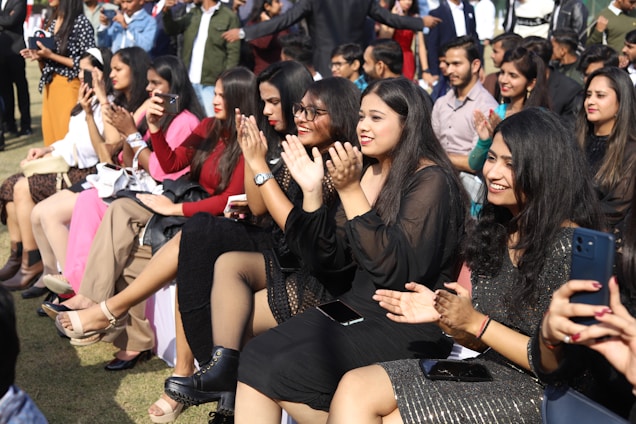 A group of people, mostly women, are seated outdoors on chairs, clapping and smiling. They are dressed in formal or semi-formal attire. The background is a mix of greenery and other people, possibly indicating a public or social event.