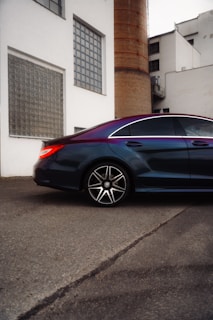 A sleek, modern car parked beside an industrial building with a large brick chimney. The car features a glossy finish and large alloy wheels, reflecting the light. The building has white walls and several rectangular windows, giving it a minimalist style.
