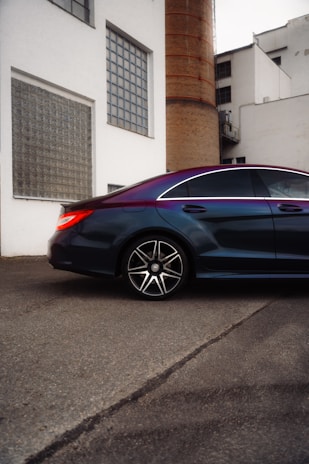A sleek, modern car parked beside an industrial building with a large brick chimney. The car features a glossy finish and large alloy wheels, reflecting the light. The building has white walls and several rectangular windows, giving it a minimalist style.