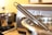 A close-up of a stainless steel whisk resting inside a metal pot, with a blurred background showing a modern kitchen setting including a stovetop and possibly a toaster.