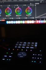 A dark setting with an illuminated control panel. The top of the image shows a computer screen displaying color wheels for video or photo editing. Below, a control surface with buttons and dials is dimly lit. The surface features a central dial with options like play, pause, fast forward, and reverse.