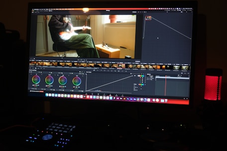 A computer monitor displays video editing software, with color grading controls and a video clip showing a person playing a guitar. The workspace includes various panels and adjustments for editing, and a speaker with a red light is visible on the desk.