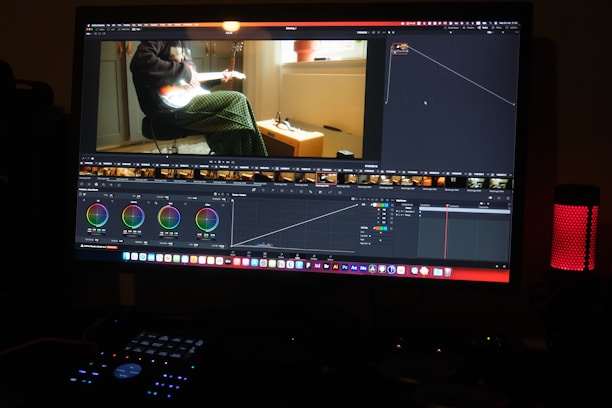 A computer monitor displays video editing software, with color grading controls and a video clip showing a person playing a guitar. The workspace includes various panels and adjustments for editing, and a speaker with a red light is visible on the desk.