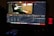 A computer monitor displays video editing software, with color grading controls and a video clip showing a person playing a guitar. The workspace includes various panels and adjustments for editing, and a speaker with a red light is visible on the desk.
