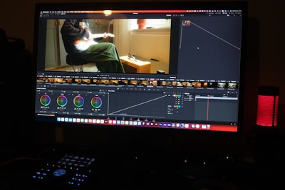 A computer monitor displays video editing software, with color grading controls and a video clip showing a person playing a guitar. The workspace includes various panels and adjustments for editing, and a speaker with a red light is visible on the desk.