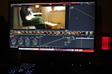 A computer screen displaying a video editing software interface. The software shows various editing tools like color wheels, a timeline with video thumbnails, and graphs. On the screen, a video clip of a person sitting and playing a guitar is visible. The workspace is dimly lit, and a red LED microphone or speaker is positioned on the desk.