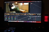 A computer screen displaying a video editing software interface. The software shows various editing tools like color wheels, a timeline with video thumbnails, and graphs. On the screen, a video clip of a person sitting and playing a guitar is visible. The workspace is dimly lit, and a red LED microphone or speaker is positioned on the desk.