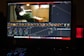 A computer screen displaying a video editing software interface. The software shows various editing tools like color wheels, a timeline with video thumbnails, and graphs. On the screen, a video clip of a person sitting and playing a guitar is visible. The workspace is dimly lit, and a red LED microphone or speaker is positioned on the desk.