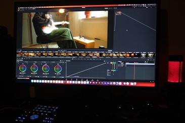 A computer screen displaying a video editing software interface. The software shows various editing tools like color wheels, a timeline with video thumbnails, and graphs. On the screen, a video clip of a person sitting and playing a guitar is visible. The workspace is dimly lit, and a red LED microphone or speaker is positioned on the desk.