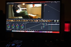 A computer screen displaying a video editing software interface. The software shows various editing tools like color wheels, a timeline with video thumbnails, and graphs. On the screen, a video clip of a person sitting and playing a guitar is visible. The workspace is dimly lit, and a red LED microphone or speaker is positioned on the desk.