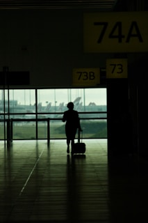 A silhouette of a person walking in an airport terminal, pulling a wheeled suitcase. Overhead signage displays gate numbers 74A, 73B, and 73. Large windows provide a view of parked airplanes and the runway outside.