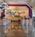 An elegant indoor event setup features a long aisle flanked by white arches adorned with vibrant floral arrangements. The seating area is arranged with chairs covered in white and peach fabric, leading up to a stage draped in golden and yellow fabrics. The stage is decorated with floral arrangements and lit candles, creating a sophisticated and warm atmosphere.