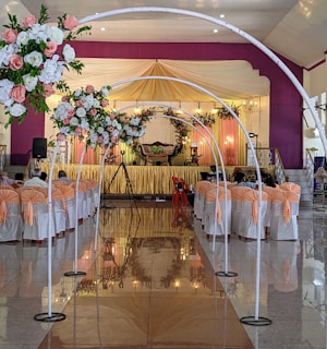 An elegant indoor event setup features a long aisle flanked by white arches adorned with vibrant floral arrangements. The seating area is arranged with chairs covered in white and peach fabric, leading up to a stage draped in golden and yellow fabrics. The stage is decorated with floral arrangements and lit candles, creating a sophisticated and warm atmosphere.