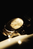 A classic wristwatch with a metallic casing and a leather strap is partially illuminated against a dark background. The watch face shows Roman numerals and the hands point to a specific time. The reflective surface of the watch catches the light, creating a warm, nostalgic glow.