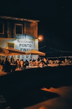 A night scene depicting an outdoor dining area by a waterfront, with several people seated at tables under large umbrellas. The establishment has a sign reading 'Restaurante Ponto Final' with a warm glow from streetlights illuminating the area. In the background, subtle lights outline a distant bridge against the night sky.