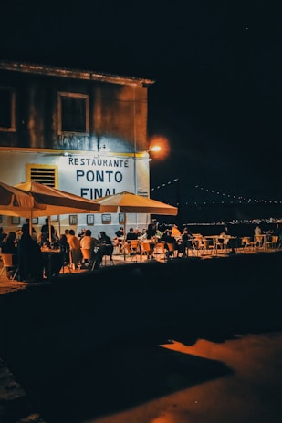 A night scene depicting an outdoor dining area by a waterfront, with several people seated at tables under large umbrellas. The establishment has a sign reading 'Restaurante Ponto Final' with a warm glow from streetlights illuminating the area. In the background, subtle lights outline a distant bridge against the night sky.