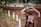 A motocross rider wearing a helmet and protective gear rides a dirt bike on a muddy trail. The rider is focused and covered in dirt, indicative of an intense race. There are white markers along the track and blurred spectators in the background.