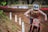 A motocross rider wearing a helmet and protective gear rides a dirt bike on a muddy trail. The rider is focused and covered in dirt, indicative of an intense race. There are white markers along the track and blurred spectators in the background.