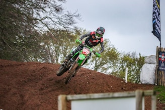 A motocross rider wearing protective gear is airborne on a dirt track, performing a jump. The rider is on a green motorbike with various decals, and the scene is set outdoors with trees in the background. A flag and some signs are visible to the right of the image.