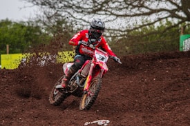 A motocross racer in a red outfit and helmet is riding through a dirt track, creating a spray of mud behind the bike. The background is filled with green foliage and trees, adding to the dynamic nature of the scene.
