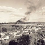 Aerial view of a residential area with houses and trees. In the distance, a large plume of dark smoke rises into the sky, contrasting with the partly cloudy weather. The landscape includes open fields and distant city buildings.