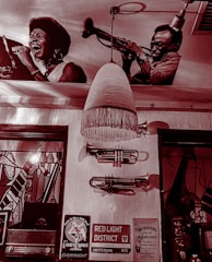 The image shows a vintage-style interior with wall decorations including large black and white photographs of musicians, specifically a singer and a trumpet player. A retro pendant light hangs from the ceiling, and there are trumpets mounted on the wall. Below, there are various signs, including one for the Red Light District in Amsterdam. The setting is reminiscent of a jazz club or music-themed venue.