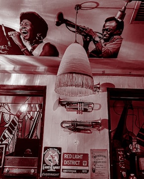 The image shows a vintage-style interior with wall decorations including large black and white photographs of musicians, specifically a singer and a trumpet player. A retro pendant light hangs from the ceiling, and there are trumpets mounted on the wall. Below, there are various signs, including one for the Red Light District in Amsterdam. The setting is reminiscent of a jazz club or music-themed venue.