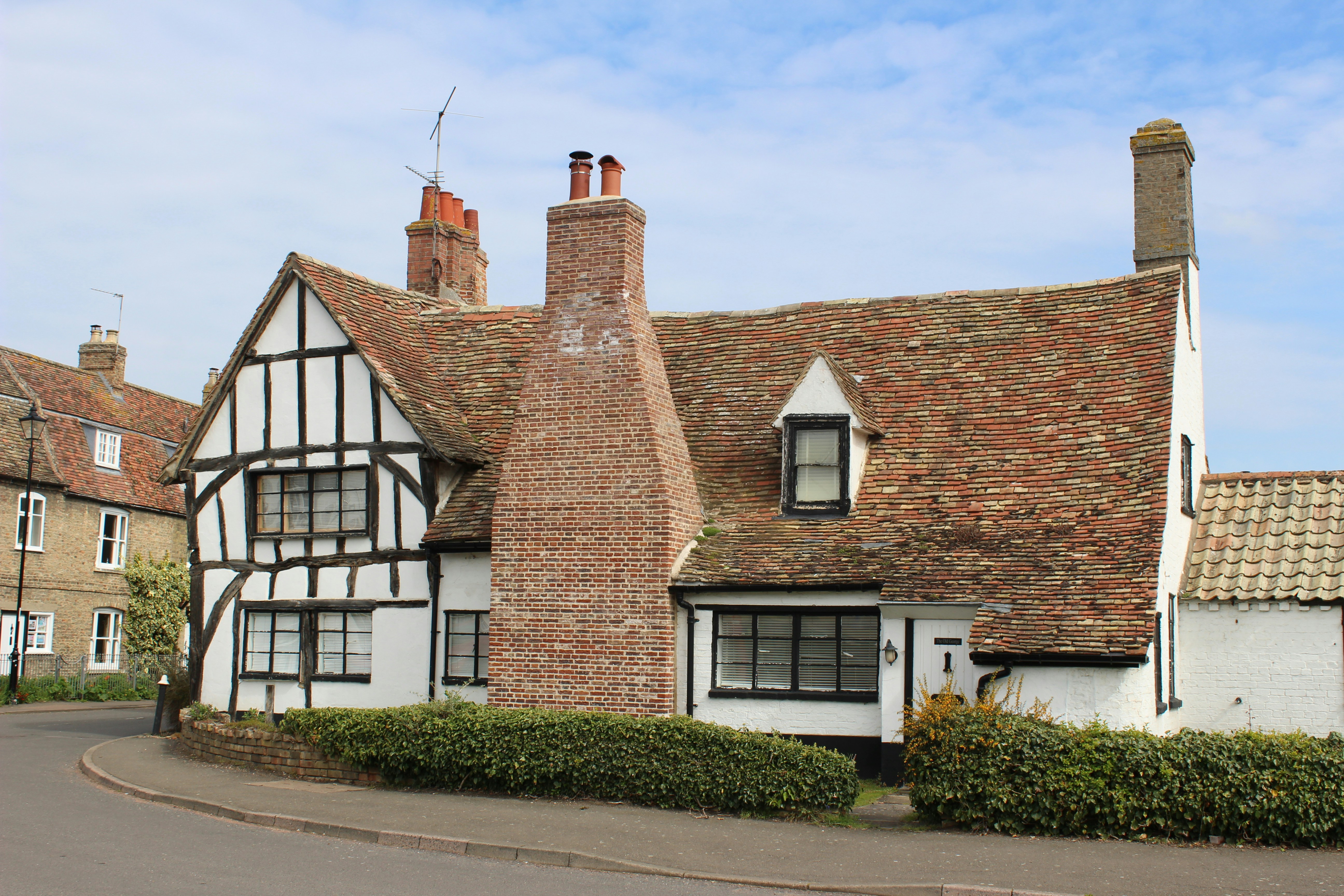 Traditional English Tudor timber-framed cottage with white walls, dark beams, and a red-tiled roof. Multiple chimneys, dormer windows, and a neat hedge on the street under a clear blue sky.