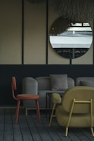 The image depicts a stylish indoor seating area with modern furniture. A round mirror hangs on a wall with a textured surface, reflecting an outdoor scene. The furniture includes an orange chair, a beige sofa with cushions, and a mustard-colored armchair, all set on wooden flooring. The decor is minimalistic with a muted and elegant color palette.