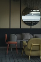 The image depicts a stylish indoor seating area with modern furniture. A round mirror hangs on a wall with a textured surface, reflecting an outdoor scene. The furniture includes an orange chair, a beige sofa with cushions, and a mustard-colored armchair, all set on wooden flooring. The decor is minimalistic with a muted and elegant color palette.