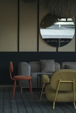 The image depicts a stylish indoor seating area with modern furniture. A round mirror hangs on a wall with a textured surface, reflecting an outdoor scene. The furniture includes an orange chair, a beige sofa with cushions, and a mustard-colored armchair, all set on wooden flooring. The decor is minimalistic with a muted and elegant color palette.